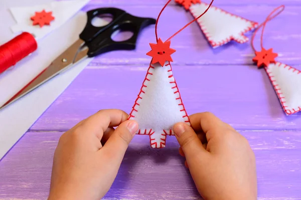 Το παιδί κατέχει μια αισθητή χριστουγεννιάτικο δέντρο διακόσμηση στα χέρια του. Το παιδί κάνει Χριστουγεννιάτικα στολίδια από τσόχα. Υλικά χειροτεχνίας και εργαλεία σε ένα ξύλινο τραπέζι. Χέρι ράψιμο μάθημα για τα παιδιά στις χειμερινές διακοπές — Φωτογραφία Αρχείου