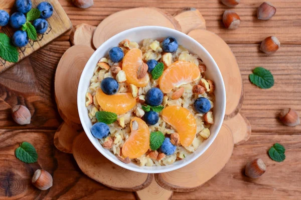 Easy oatmeal with citrus, berries and nuts. Porridge with tangerines, blueberries, hazelnuts and mint in a white bowl and on a wooden table. Detox diet. Maintaining a healthy lifestyle. Top view