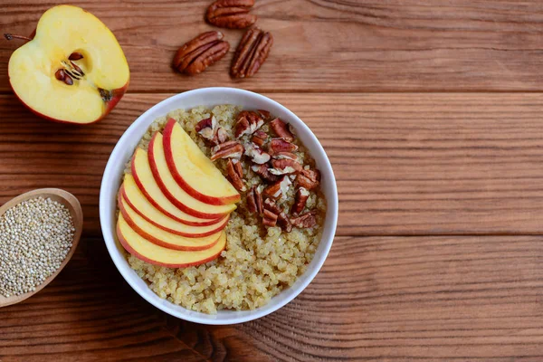 Quinoa Breakfast porridge. Vegetarian quinoa porridge with fresh apples and pecan nuts in a white bowl and on wooden background with copy space for text. Rustic style. Top view