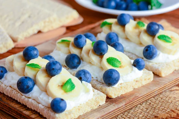 Soft cheese cream, bananas and berries sandwiches. Cracker bread with toppings on wooden board. Closeup