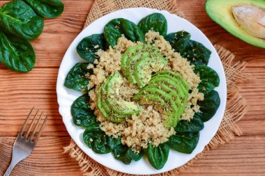 Quinoa, avokado ve taze ıspanak salatası bir beyaz plaka ve kahverengi ahşap zemin. Sebzeli quinoa avokado kahvaltı fikir. Üstten Görünüm