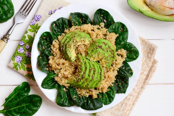 Quinoa porridge with fresh spinach and avocado slices. Vegan quinoa spinach avocado salad on a white plate. White wooden background. Top view. Quinoa avocado breakfast bowl. Avocado quinoa recipe. Quinoa avocado spinach salad