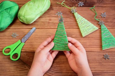 Çocuk elinde bir Noel ağacı süslemesi tutuyordu. Çocuk Noel ağacı desenli yün ipliği gösteriyor. Çocuklar için mutlu noel ağacı projesi. Ahşap bir masada yeşil pamuk ipliği. Şık Noel ağacı süsleme fikri