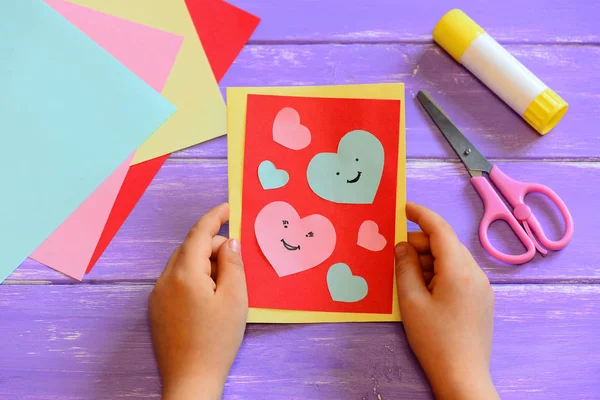Child is holding a Valentines day card diy in his hands. Small kid made a handmade Valentines day greeting card with hearts smiles. Cute and simple paper crafts for kindergarten. Paper cutting hearts. Closeup. Easy paper crafts for kids at home