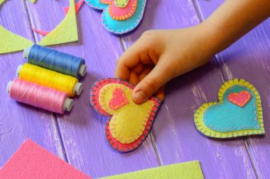 Çocuk bir keçe kalp valentine elinde tutuyor. Çocuk bir keçe kalp valentine yaptı. Renkli Keçe Kalpler, makas, iş parçacığı üzerinde ahşap bir masa ayarla. Sevgililer günü el sanatları kavramı. Çocuklar için eğlenceli dikiş projeler. Ev yapımı Sevgililer günü hediye fikri. Kalp el sanatları