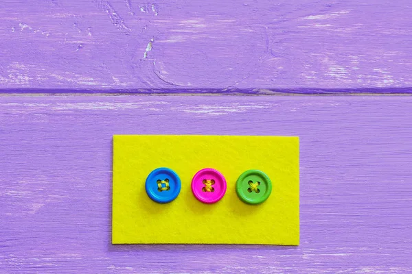 Various ways to sew buttons to felt. Yellow flat felt with bright buttons isolated on wooden background with copy space. Sewing project. How to sew a button by hand. Decoration idea. Decorating clothes with buttons. Button design. Button crafts diy