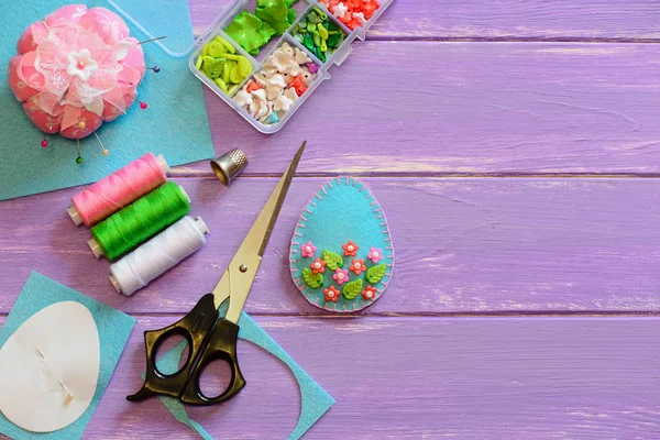 Nice Easter egg ornament with plastic flowers and leaves. Felt egg crafts, paper template, scissors on the wooden background with copy space. Quick Easter crafts. Home sewing. Sewing concept workplace. Tailor workplace. Hand sewing concept workplace