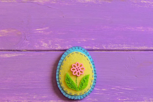 Felt Easter egg ornament with pink flower button isolated on a wooden background with copy space. Fun Easter sewing crafts for children. Happy Easter greeting card. Spring holiday. Patchwork felt Easter egg. Easter sewing projects. Easter crafts