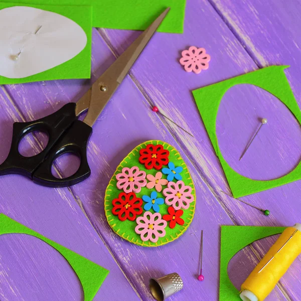 Felt Easter egg decoration idea. Hodemade felt Easter egg with colored wooden flower buttons. Felt scrap, scissors, thimble, thread, paper template, pins on the table. Holiday crafts. Craft projects. Easter craft ideas. Easter spring crafts