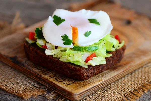 Delicious poached egg sandwich. Simple poached egg on rye bread slice with fresh vegetable mix and parsley. Healthy breakfast idea. Rustic wooden background. Closeup. Cheap healthy sandwich. Vegetarian sandwich idea
