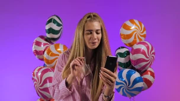 Kulaklıklı, akıllı telefonlu bir kız, bir şarkı söylüyor, mor renkli balonlarla, şekerlerin arka planında — Stok video