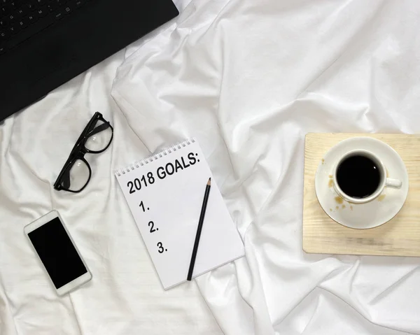Top view of bed, notepad with black inscription 2018 goals, pencil, notebook, smart phone, glasses, white coffee cup on wood board. Future new year plans checklist mock up with copy space