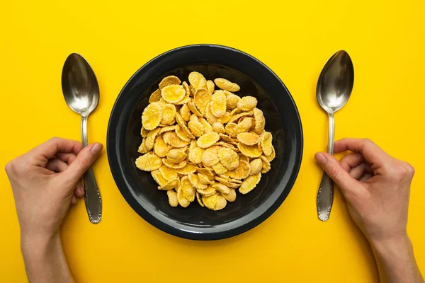 Sarı bir arka plan üzerinde siyah bir tabak içinde sarı mısır gevreği. Eller bir kaşıkla yemek her iki el ile. — Stok fotoğraf