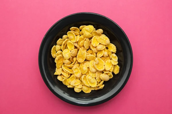 Yellow corn flakes in a black plate on a pink background.