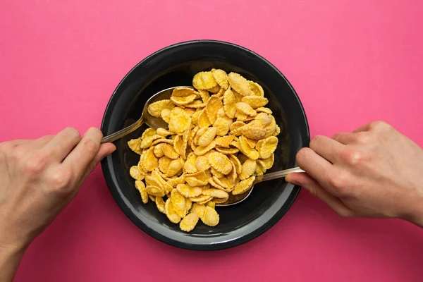 Flocons de maïs jaune dans une assiette noire sur un fond rose. Mains avec une cuillère, manger avec les deux mains . — Photo