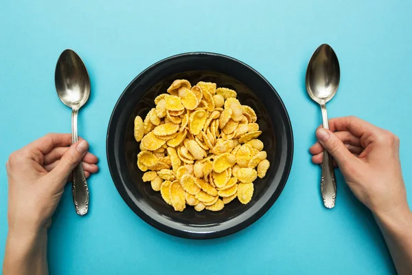 Gele cornflakes in een zwarte plaat op een blauwe achtergrond. Handen met een lepel, eten met beide handen. — Stockfoto