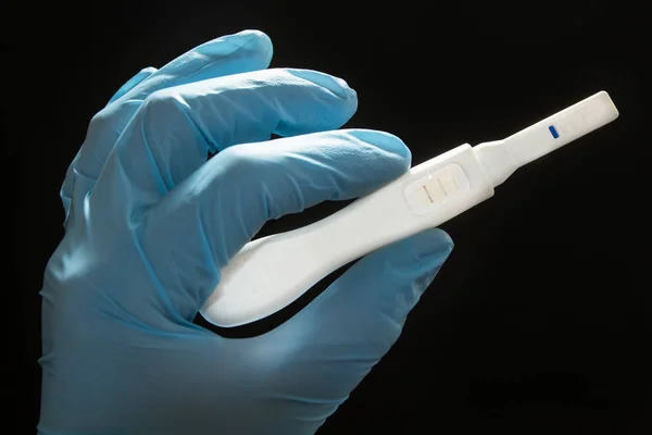 Positive pregnancy test with two strips in the hand in a blue medical glove on a black background. Artificial insemination, concept. — Stock Photo, Image