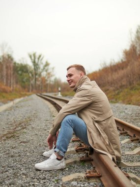 Sonbahar havası, yeraltında yağmurluk giymiş genç bir adam, sokak tarzı. Trençkotlu, spor ayakkabılı ve kot pantolonlu genç bir adam demiryolu raylarında oturuyor..