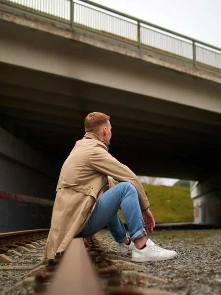 Autumn mood, young guy in a raincoat trench coat underground, street style. A young guy in a trench coat, sneakers and jeans sits on a rail of railway tracks. Stock Image