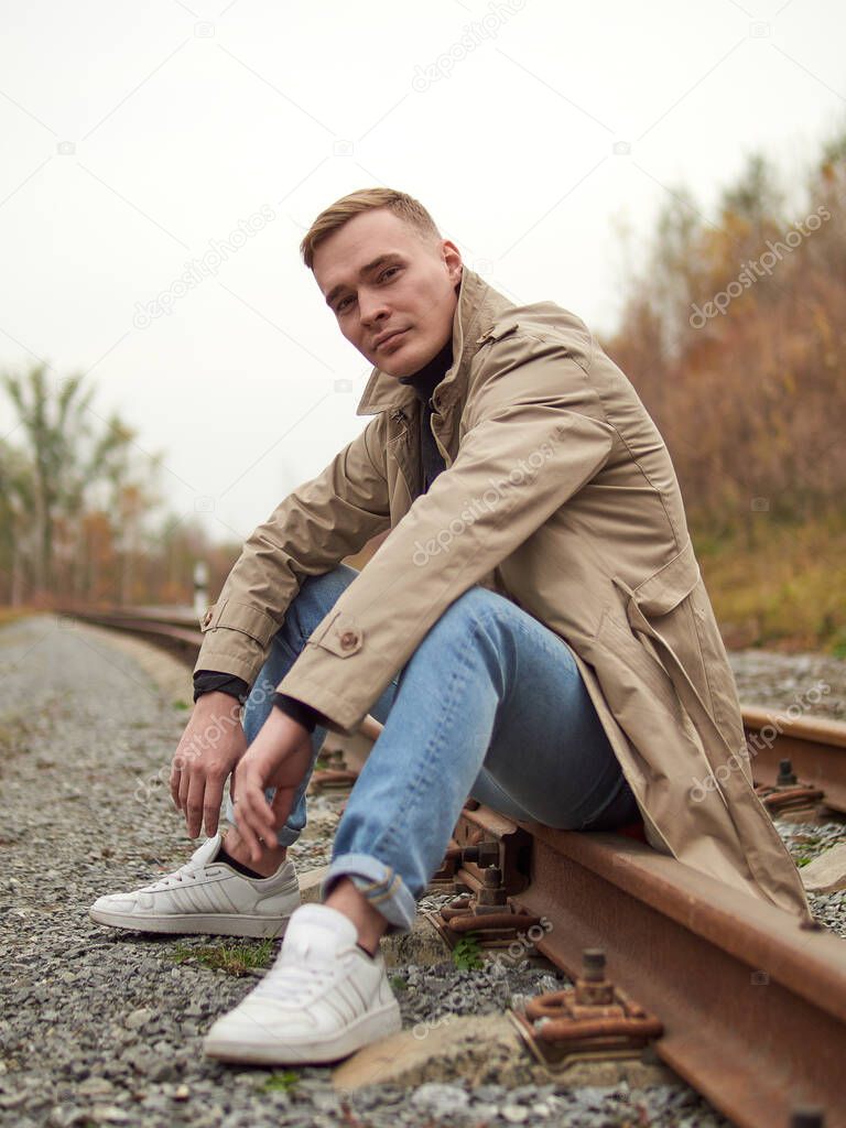 Autumn mood, young guy in a raincoat trench coat underground, street style. A young guy in a trench coat, sneakers and jeans sits on a rail of railway tracks.