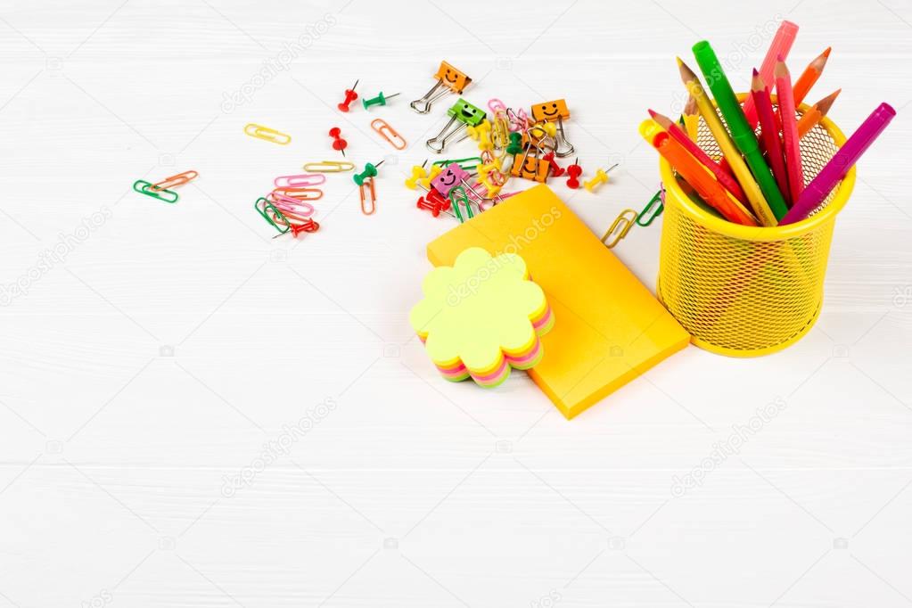 Colorful pencils and felt-tip pens in yellow pencil case, colorful notepapers, paper clips and stationery nails on white wooden background