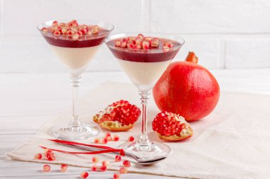 Creamy vanilla panna cotta with red jelly in beautiful glasses, fresh ripe pomegranate on white wooden background. clipart