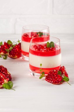 Güzel gözlük kırmızı jöle, Beyaz ahşap zemin üzerinde taze olgun Nar ile kremsi vanilya panna cotta.