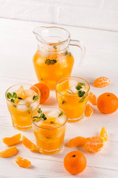 Cocktail al mandarino con ghiaccio e menta in bellissimi bicchieri e brocca, agrumi freschi maturi su fondo bianco in legno. Succo d'arancia dolce. Chiudi la fotografia. Focus selettivo — Foto Stock