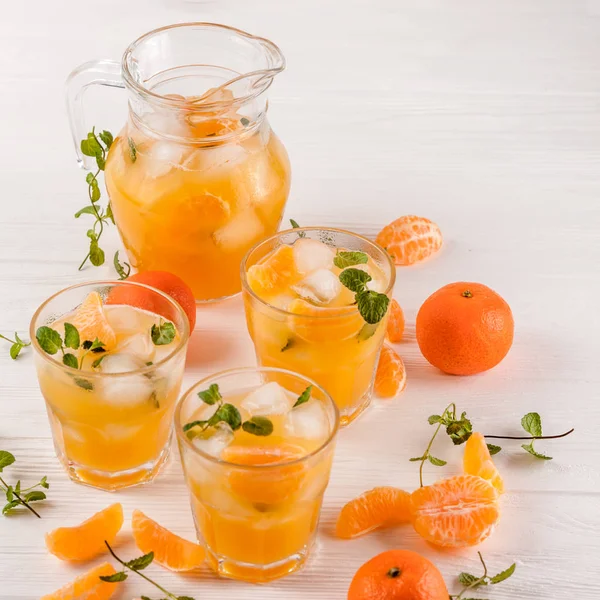 Cocktail al mandarino con ghiaccio e menta in bellissimi bicchieri e brocca, agrumi freschi maturi su fondo bianco in legno. Succo d'arancia dolce. Chiudi la fotografia. Focus selettivo — Foto Stock