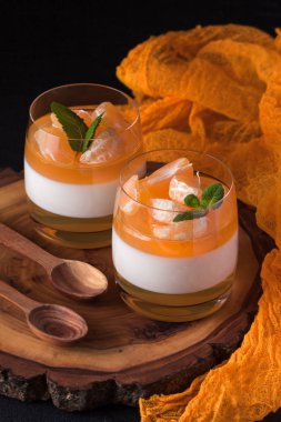 Kremsi panna cotta ile güzel bardaklarda portakal jöle, taze olgun mandarin siyah arka plan üzerine ahşap bölümünde. Lezzetli İtalyan tatlı. Modern karanlık ruh stili.