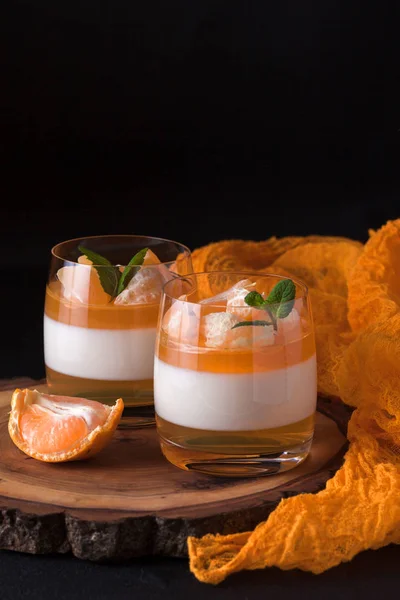 Smetanové panna cotta s oranžové želé v krásné brýle, čerstvé zralé mandarinka na dřevěnou desku na černém pozadí. Italský zákusek. Styl moderní ponurá nálada. — Stock fotografie