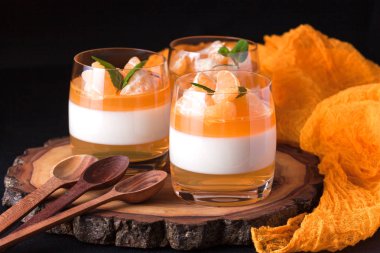 Kremsi panna cotta ile güzel bardaklarda portakal jöle, taze olgun mandarin siyah arka plan üzerine ahşap bölümünde. Lezzetli İtalyan tatlı. Modern karanlık ruh stili.