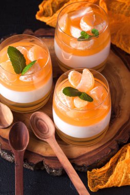 Kremsi panna cotta ile güzel bardaklarda portakal jöle, taze olgun mandarin siyah arka plan üzerine ahşap bölümünde. Lezzetli İtalyan tatlı. Modern karanlık ruh stili.