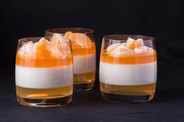 Kremsi panna cotta ile güzel bardaklarda portakal jöle, taze olgun mandarin siyah arka plan üzerine ahşap bölümünde. Lezzetli İtalyan tatlı. Modern karanlık ruh stili.