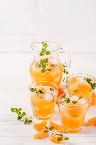 Cocktail al mandarino con ghiaccio e menta in bellissimi bicchieri e brocca, agrumi freschi maturi su fondo bianco in legno. Succo d'arancia dolce. Chiudi la fotografia. Focus selettivo — Foto Stock