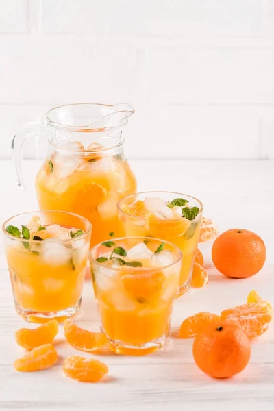 Cocktail al mandarino con ghiaccio e menta in bellissimi bicchieri e brocca, agrumi freschi maturi su fondo bianco in legno. Succo d'arancia dolce. Chiudi la fotografia. Focus selettivo — Foto Stock