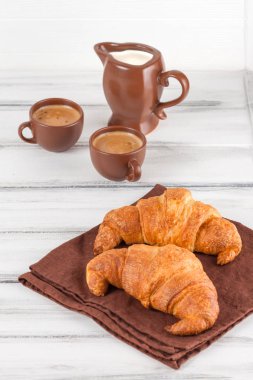 Taze pişmiş kruvasanlar üzerinde kahverengi peçete, krem, kahve Beyaz ahşap zemin üzerinde seramik yemekler için. Kahvaltı için taze hamur işleri. Lezzetli tatlı. Portre fotoğrafçılığı. Dikey başlık