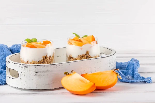 Persimmon creamy trifle in beautiful glasses, fresh ripe fruit slices on white wooden background. Healthy vegetarian food. Delicious dessert. Close up photography. Selective focus. — Stock Photo, Image