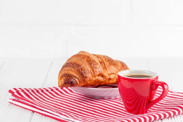 Croissant appena sfornato su tovagliolo, tazza di caffè in tazza rossa su fondo di legno bianco. Colazione francese. Pasticcini freschi per colazione. Delizioso dessert. Fotografia da vicino. Bandiera orizzontale — Foto Stock