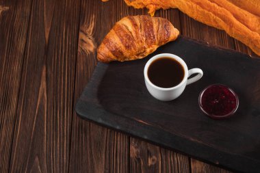 Kruvasan reçel, kahverengi ahşap zemin üzerinde beyaz fincan kahve fırından yeni çıkmış. Fransız kahvaltı. Kahvaltı için taze hamur işleri. Modern karanlık ruh tarzı. Lezzetli tatlı. Yatay başlık