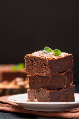 Çikolatalı kek kare parçalar halinde ceviz, beyaz plaka üzerine nane yaprakları ve siyah arka plan üzerine kakao ile dekore edilmiştir. Lezzetli tatlı. Karanlık ruh. Fotoğraf kadar kapatın. Seçici odak