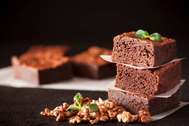 Çikolatalı kek kare parçalar halinde ceviz, beyaz plaka üzerine nane yaprakları ve siyah arka plan üzerine kakao ile dekore edilmiştir. Lezzetli tatlı. Karanlık ruh. Fotoğraf kadar kapatın. Seçici odak