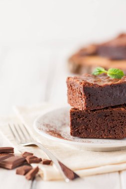 Çikolatalı kek kare parçalar halinde beyaz plaka üzerine nane yaprakları ve beyaz vintage ahşap zemin üzerine kakao tozu ile dekore edilmiştir. Amerikan geleneksel lezzetli tatlı. Fotoğraf kadar kapatın