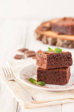 Çikolatalı kek kare parçalar halinde beyaz plaka üzerine nane yaprakları ve beyaz vintage ahşap zemin üzerine kakao tozu ile dekore edilmiştir. Amerikan geleneksel lezzetli tatlı. Fotoğraf kadar kapatın