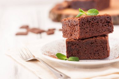 Çikolatalı kek kare parçalar halinde beyaz plaka üzerine nane yaprakları ve beyaz vintage ahşap zemin üzerine kakao tozu ile dekore edilmiştir. Amerikan geleneksel lezzetli tatlı. Fotoğraf kadar kapatın