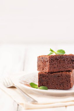 Çikolatalı kek kare parçalar halinde beyaz plaka üzerine nane yaprakları ve beyaz vintage ahşap zemin üzerine kakao tozu ile dekore edilmiştir. Amerikan geleneksel lezzetli tatlı. Fotoğraf kadar kapatın
