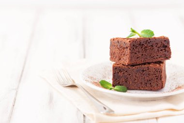 Çikolatalı kek kare parçalar halinde beyaz plaka üzerine nane yaprakları ve beyaz vintage ahşap zemin üzerine kakao tozu ile dekore edilmiştir. Amerikan geleneksel lezzetli tatlı. Fotoğraf kadar kapatın
