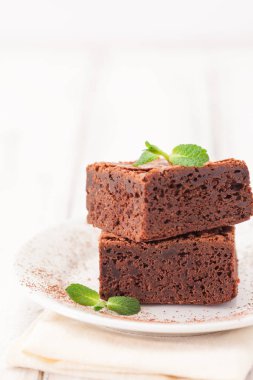 Çikolatalı kek kare parçalar halinde beyaz plaka üzerine nane yaprakları ve beyaz vintage ahşap zemin üzerine kakao tozu ile dekore edilmiştir. Amerikan geleneksel lezzetli tatlı. Fotoğraf kadar kapatın