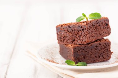 Çikolatalı kek kare parçalar halinde beyaz plaka üzerine nane yaprakları ve beyaz vintage ahşap zemin üzerine kakao tozu ile dekore edilmiştir. Amerikan geleneksel lezzetli tatlı. Fotoğraf kadar kapatın
