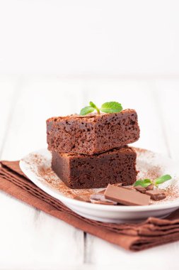 Çikolatalı kek kare parçalar halinde beyaz plaka üzerine nane yaprakları ve beyaz vintage ahşap zemin üzerine kakao tozu ile dekore edilmiştir. Amerikan geleneksel lezzetli tatlı. Fotoğraf kadar kapatın